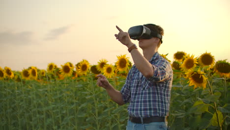 A-young-farmer-in-plaid-shirt-and-jeans-uses-VR-glasses-on-the-field-with-sunflowers-for-scientific-development.-These-are-modern-technologies-in-summer-evening.